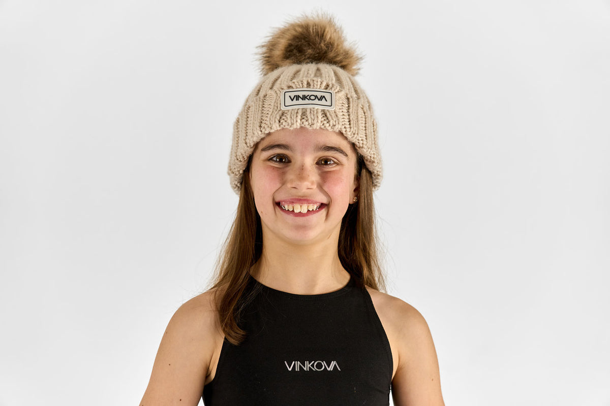 Berretto invernale Vinkova in maglia beige con pompon, indossato da una ginnasta.